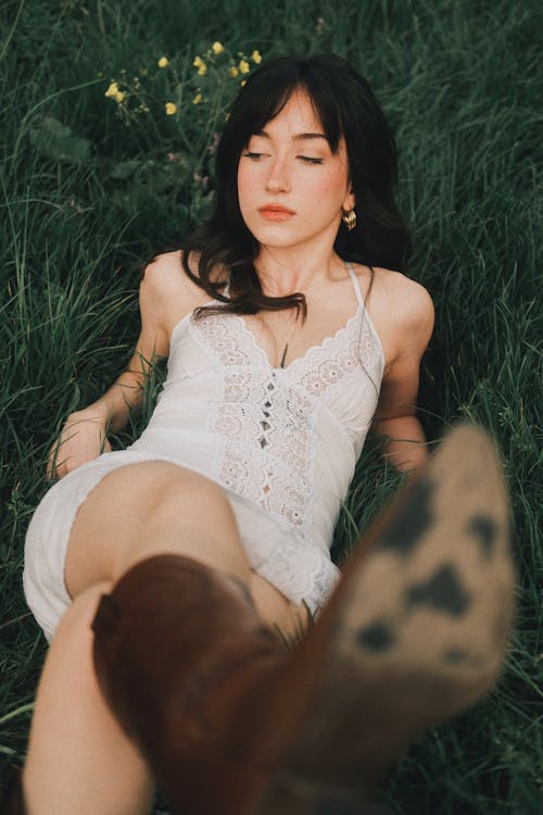 A woman laying in the grass wearing a white dress and cowboy boots