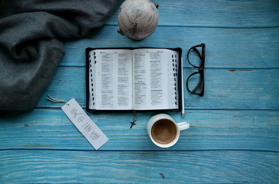 An open Bible with a bookmark, coffee, glasses, and a globe on a blue wooden table.
