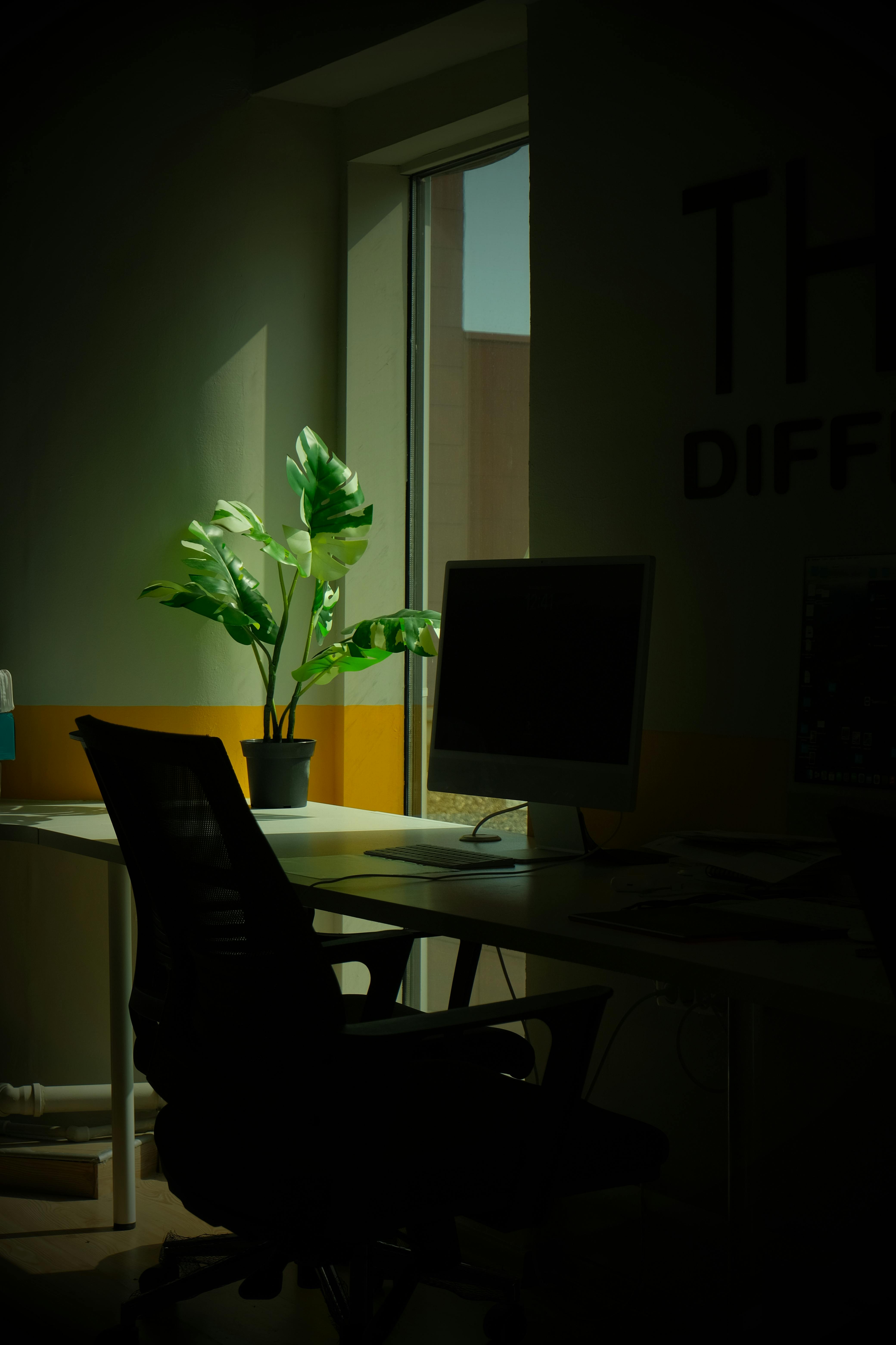 Un espacio de oficina minimalista con escritorio, monitor de computadora y planta en maceta cerca de una ventana.