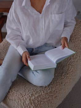A woman wearing casual clothes sits indoors, reading a book in a relaxed setting.
