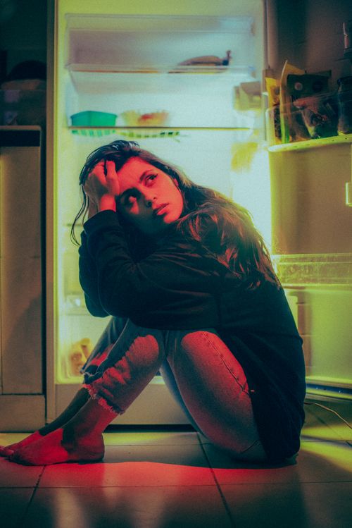 Free A contemplative woman with long hair sits by an open fridge, bathed in warm light. Stock Photo