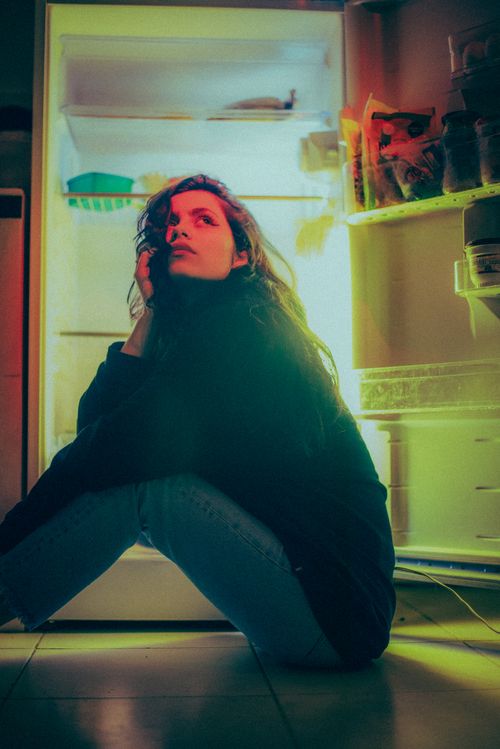 Free Young woman sits in front of open fridge at night, lost in thought. Stock Photo