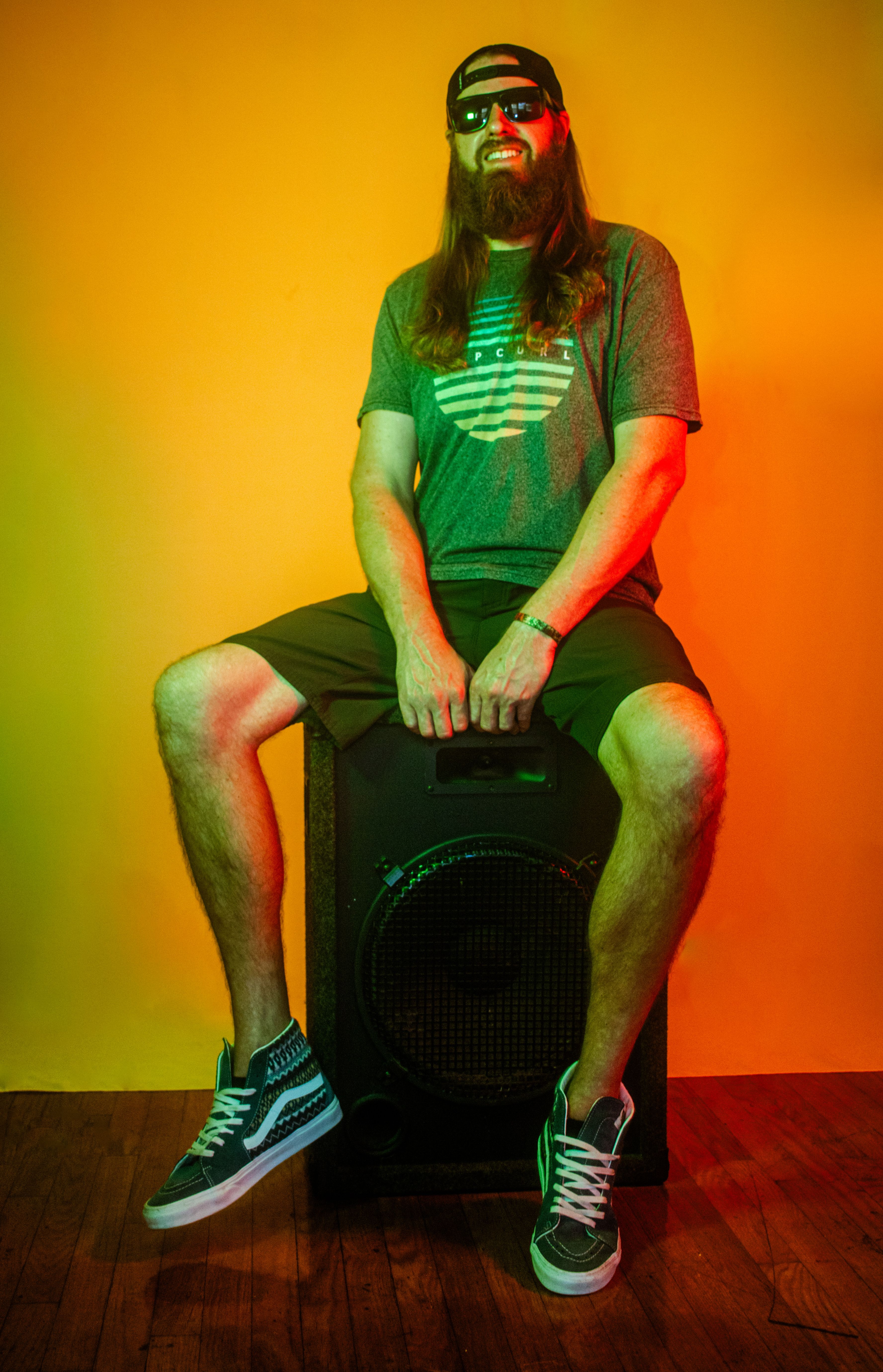 A stylish musician with sunglasses and long hair poses on a speaker in a vibrant studio.