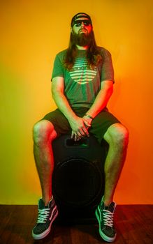 Bearded man wearing cap and sunglasses poses seated on a speaker in vibrant lighting.