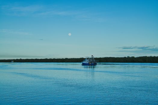A tranquil river view at dawn featuring a lone fishing boat and a moonlit sky.