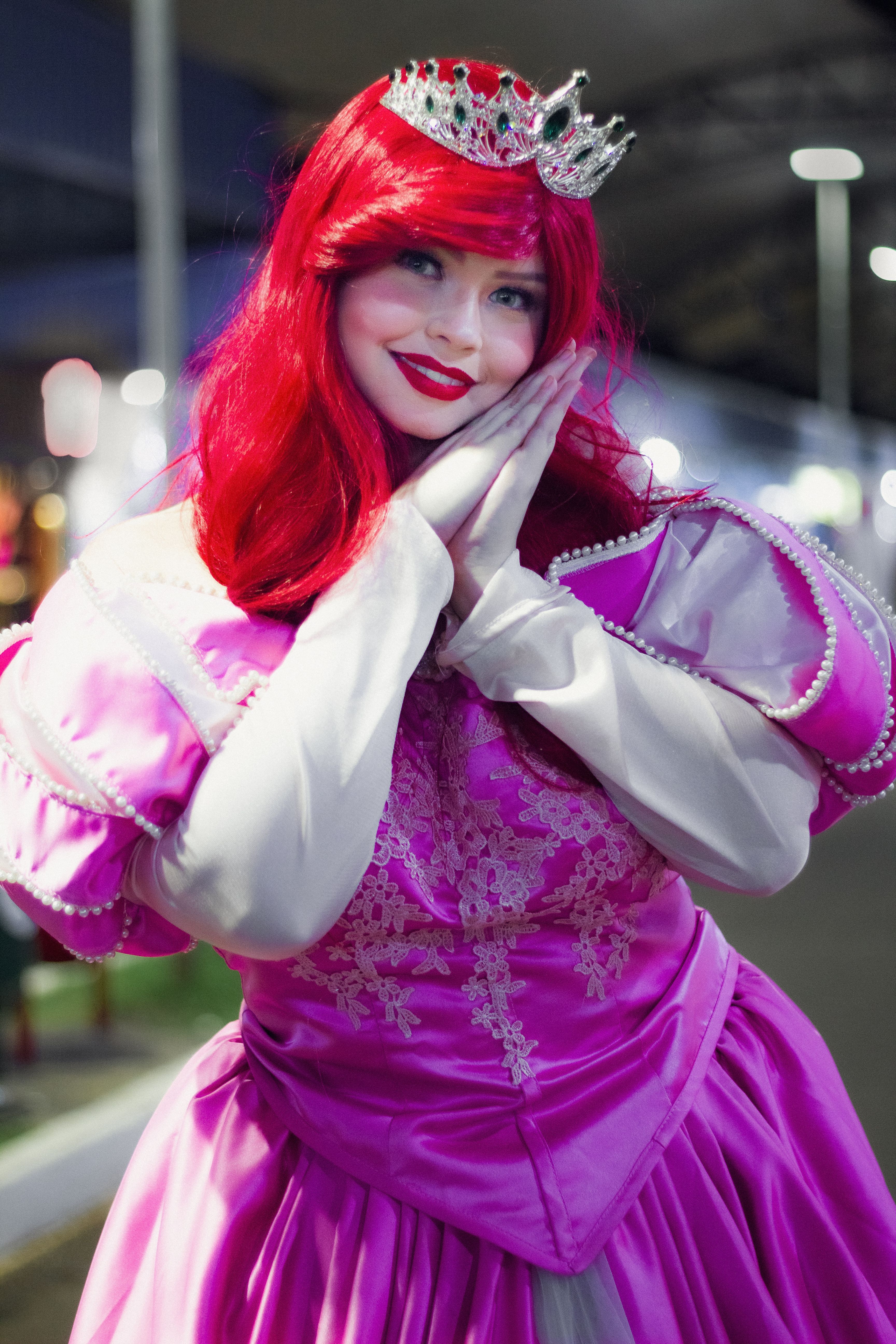 A vibrant cosplay photo of a woman in a pink gown smiling and posing outdoors at night.