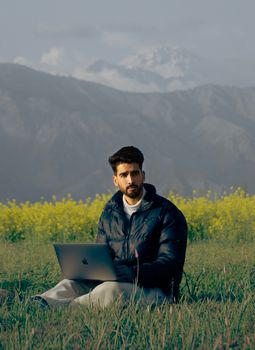 A man sits with a laptop in a lush meadow with mountain backdrop, perfect for remote work inspiration.
