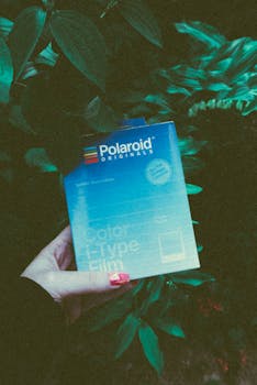 A hand holding Polaroid I-Type film pack against a backdrop of green foliage.