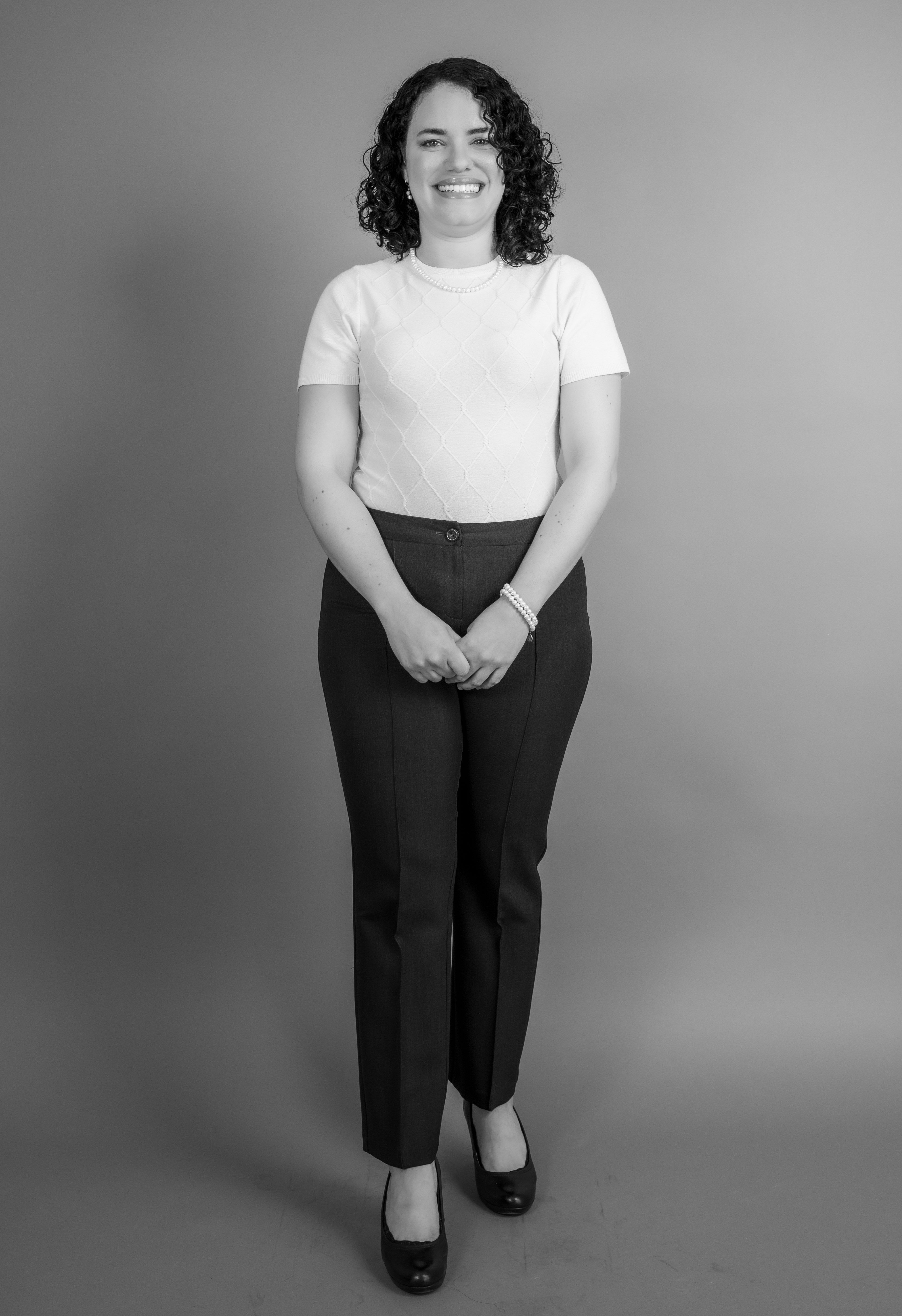 A black and white portrait of a smiling woman standing in a studio, wearing a t-shirt and pants.
