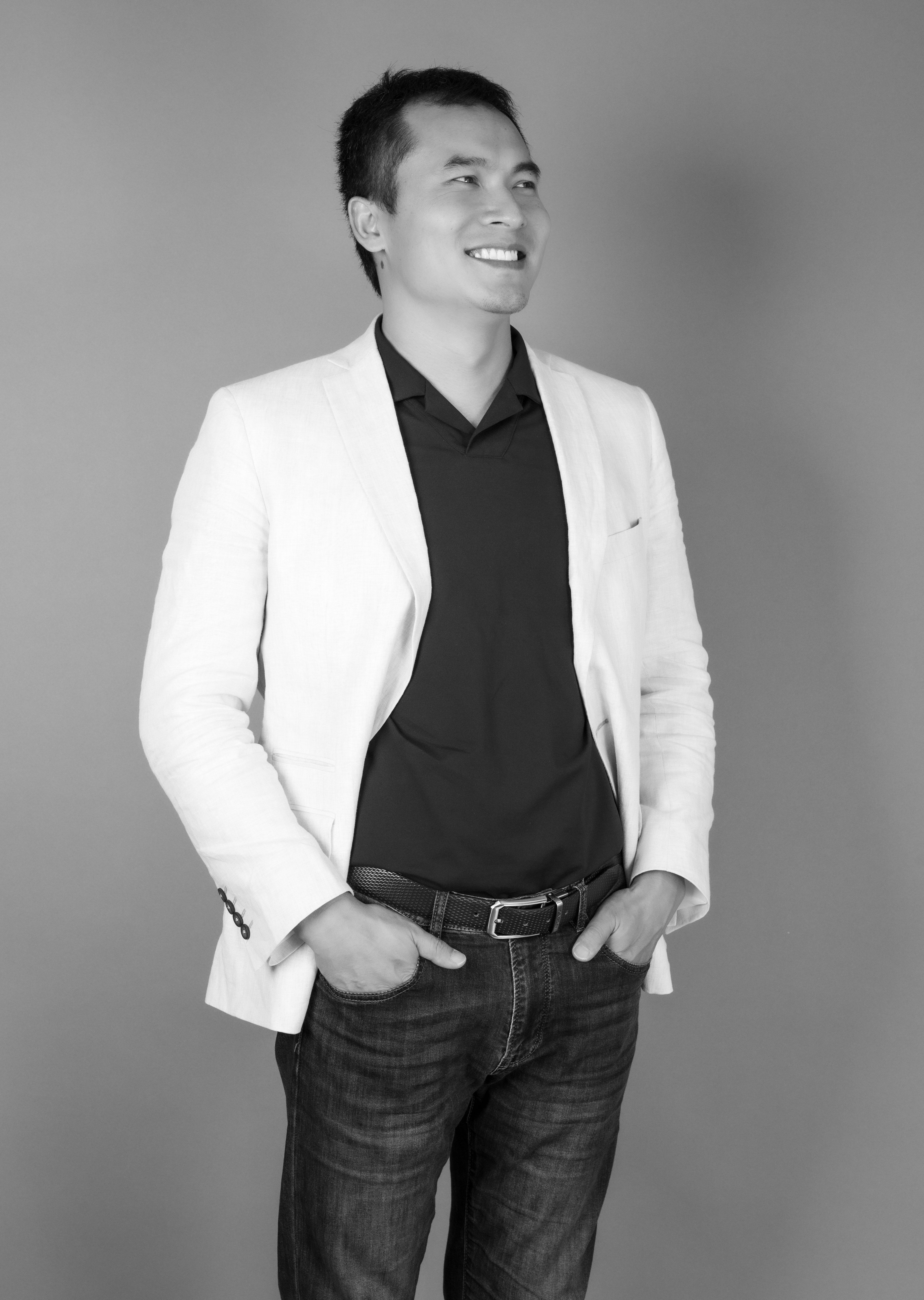 Black and white portrait of a smiling man in casual fashion attire with hands in pockets.