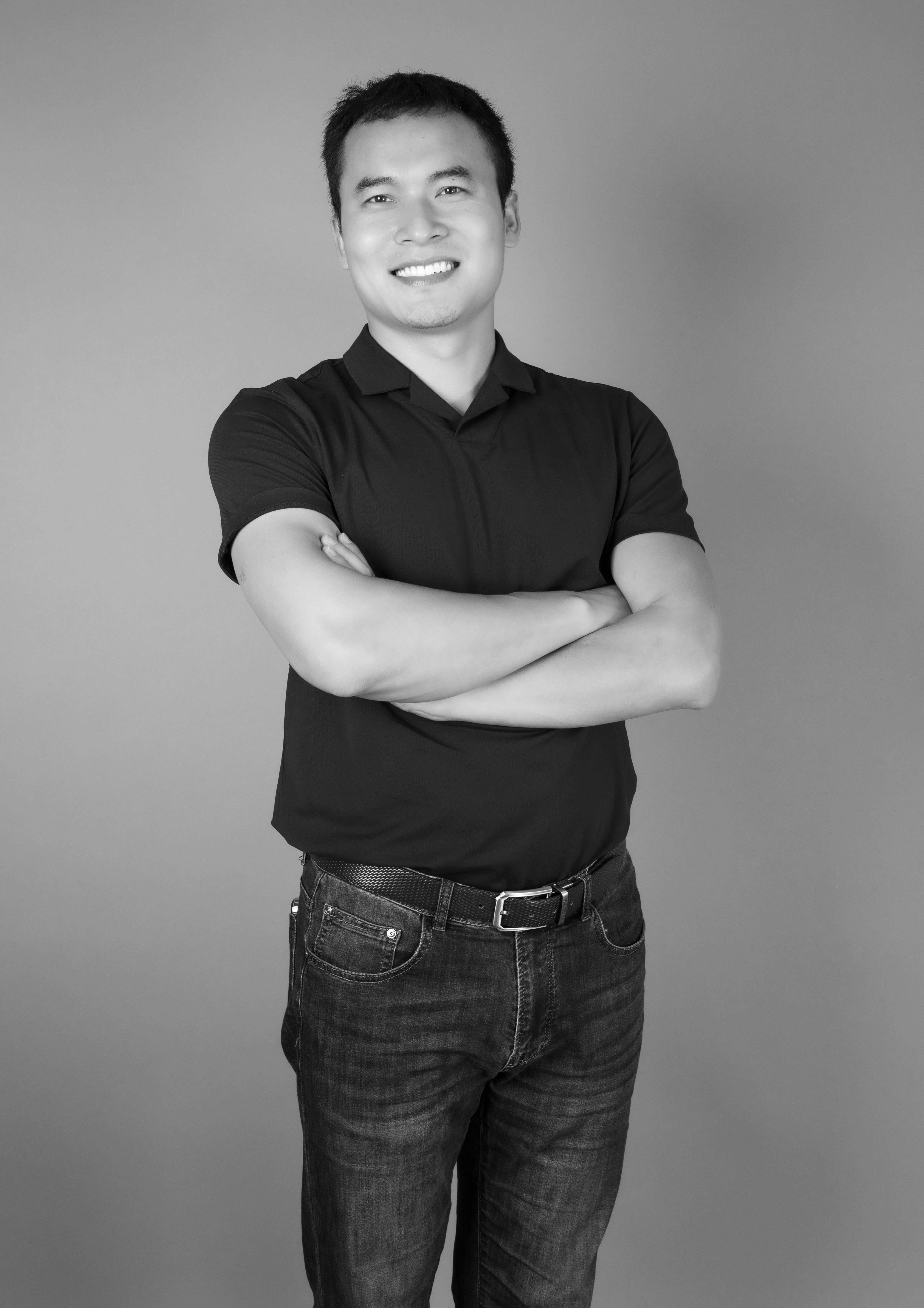 Black and white portrait of a smiling Asian man in casual attire standing with crossed arms.