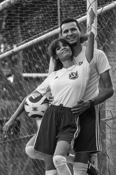 Couple of soccer players in uniforms smiling and posing by a goal post.