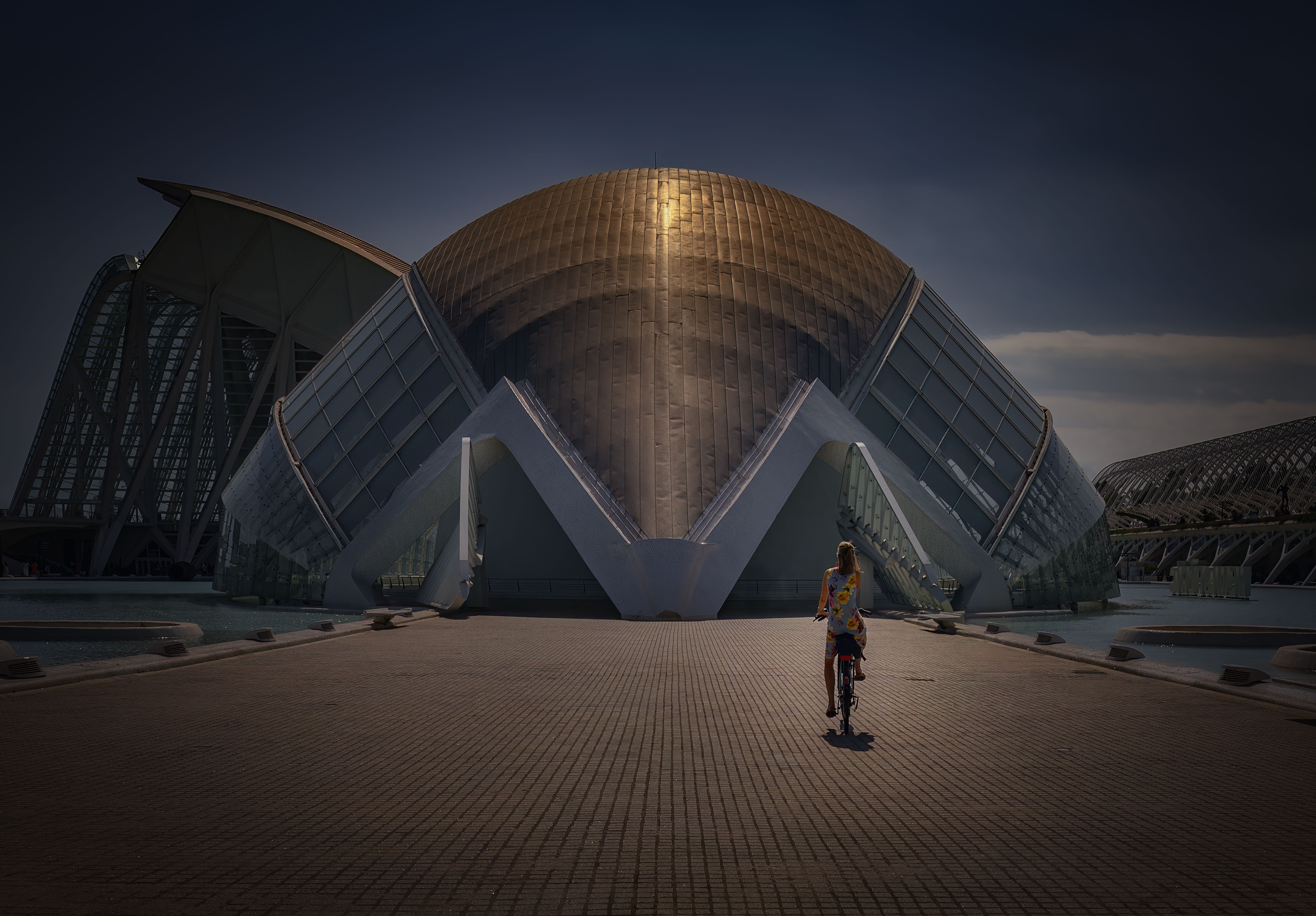 Dramatic view of hemispherical dome at City of Arts and Sciences, Valencia. A woman rides a bicycle towards it.