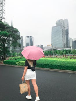 雨天，一位打着粉红色伞的妇女在城市公园散步，背景是摩天大楼