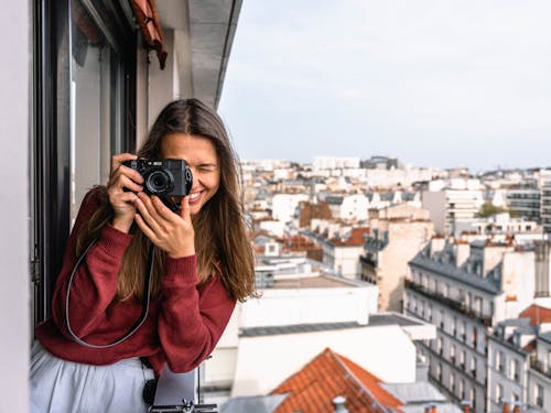 Mujer Vistiendo Suéter Granate De Pie En La Terraza Con Cámara Mientras Sonríe Con Vistas A Casas Y Edificios