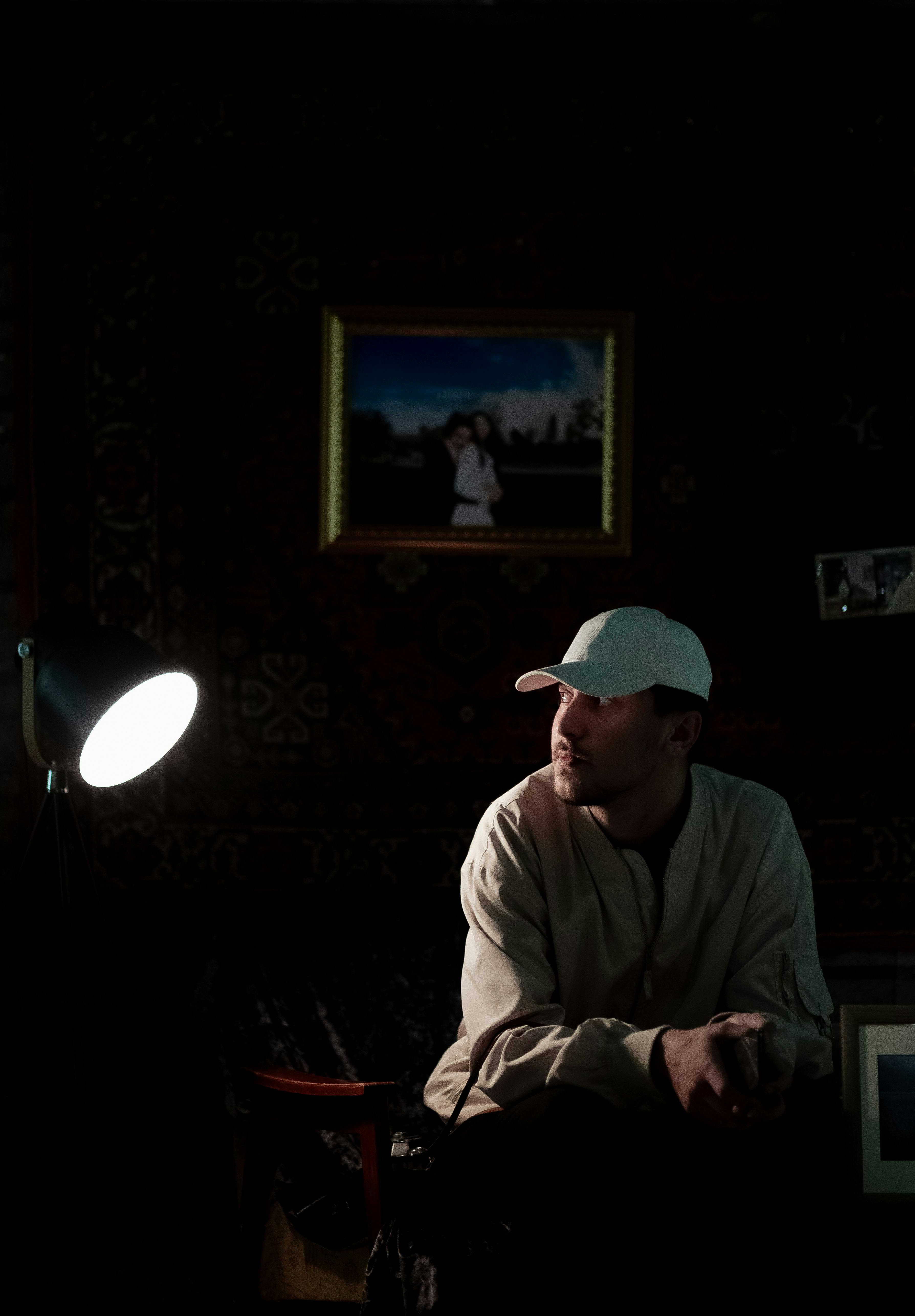 Man in a cap sitting in dimly lit room with dramatic lighting and framed photos.