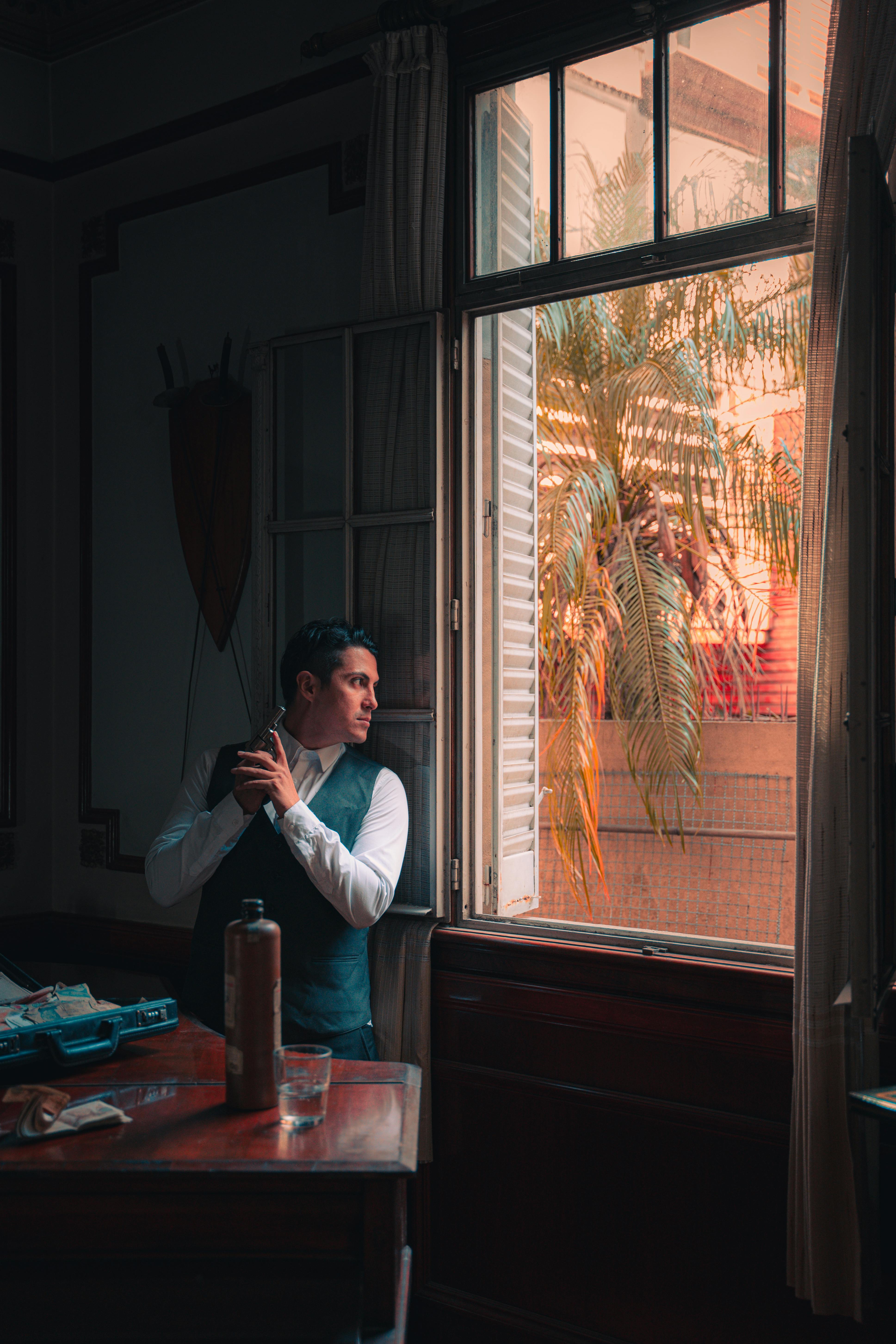 A man in a formal vest holds a gun while looking outside through a window, creating a mysterious ambiance.