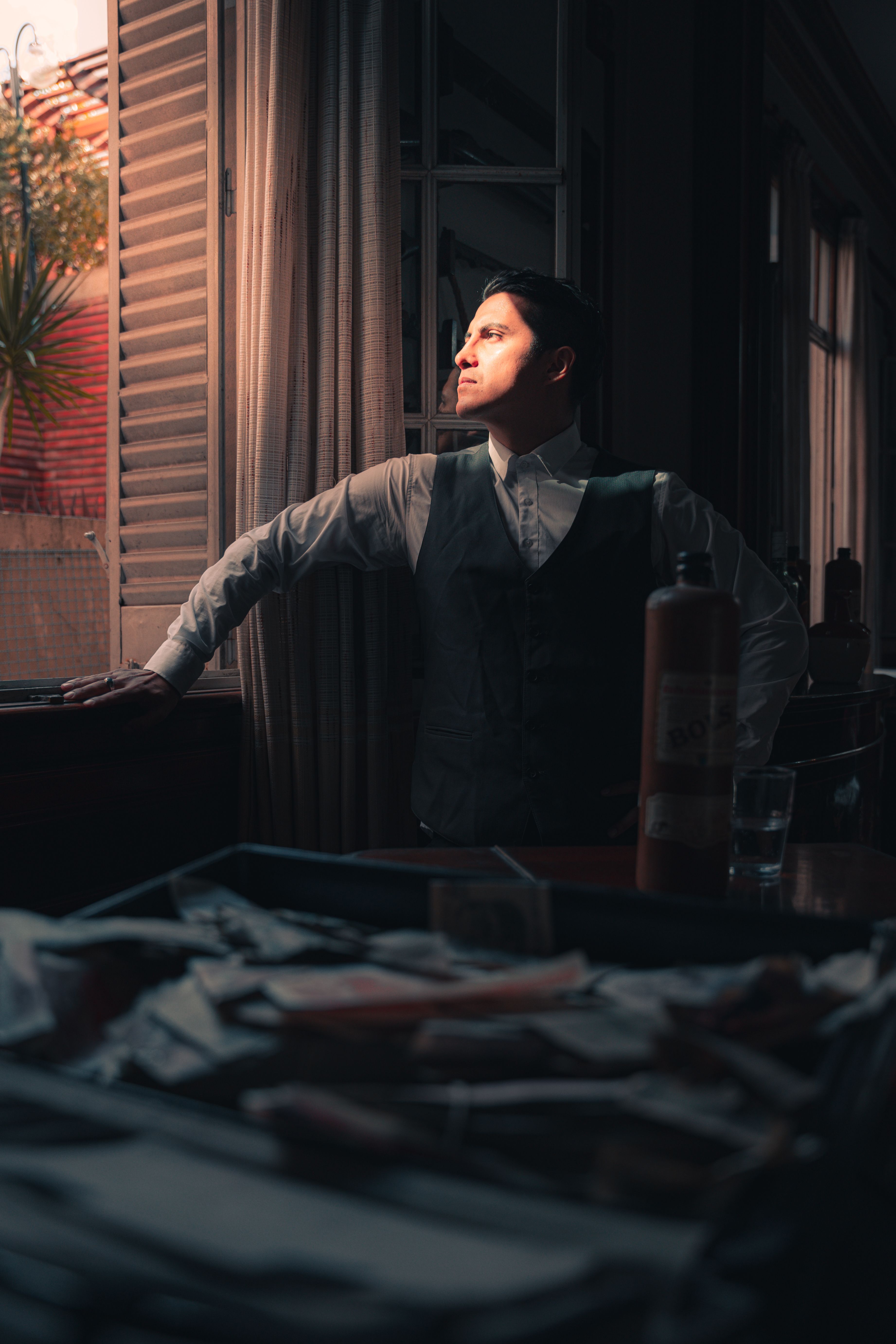 A man in vintage attire stands pensively indoors, illuminated by warm evening light.