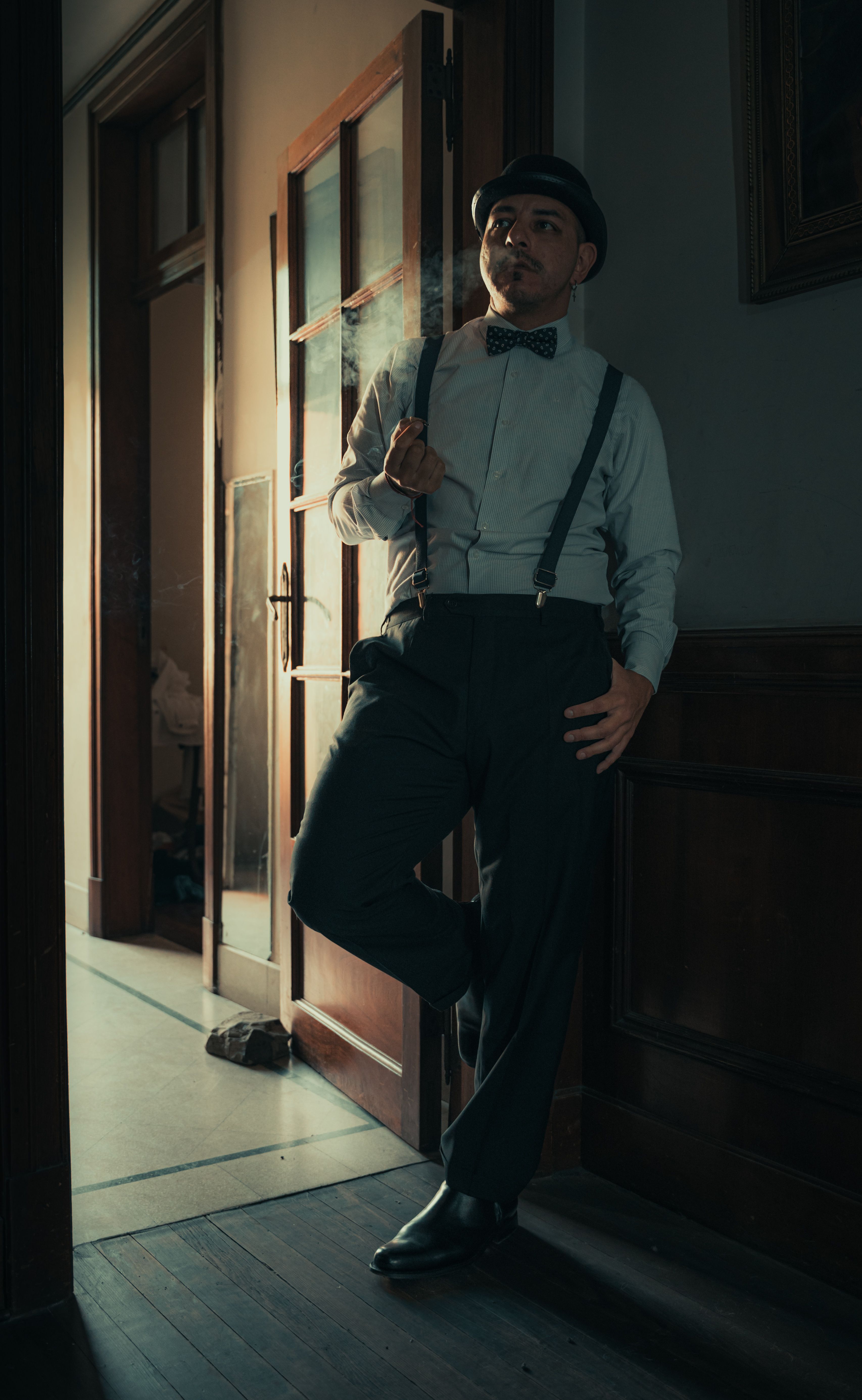 Vintage fashion portrait of a man smoking in 1920s attire with bow-tie and suspenders indoors.