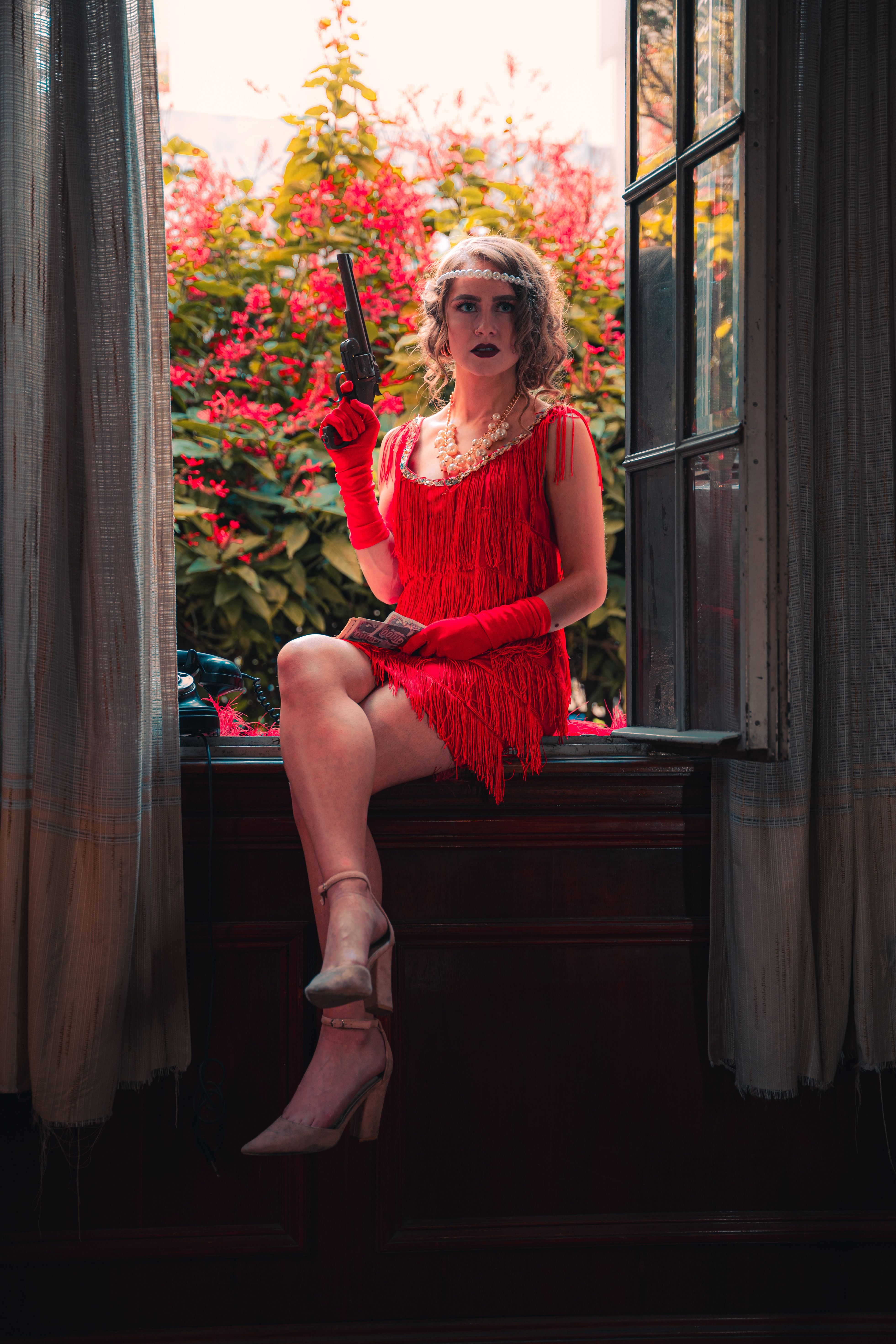 A striking vintage-style portrait of a woman in red attire holding a revolver, set indoors by a window.