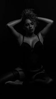 Sensual black and white portrait of a woman in lingerie sitting cross-legged in a dark setting.