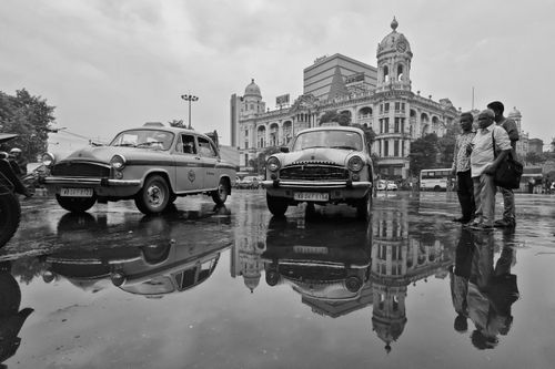 Free Black and white scene of vintage taxis and people with Kolkata's iconic architecture reflected in water. Stock Photo