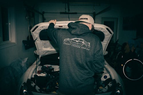 A man in a hoodie looking at the hood of a car