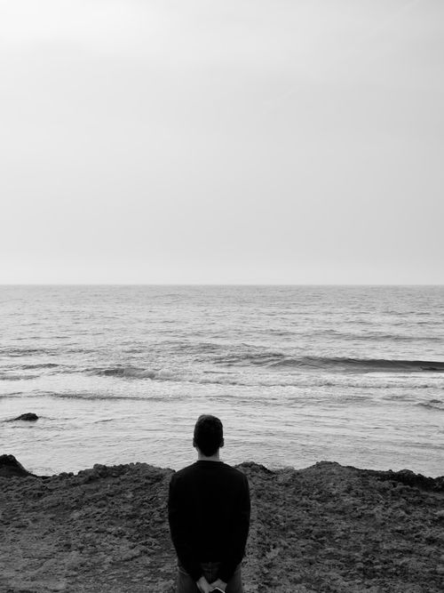 Free A man standing on the shore, gazing at the vast, serene sea in black and white. Stock Photo