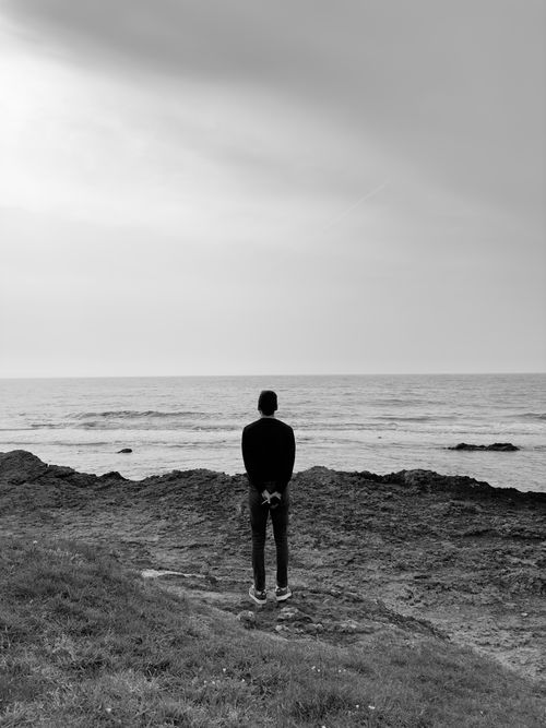 Free A man stands on a rocky shore, gazing out at the sea, captured in black and white. Stock Photo