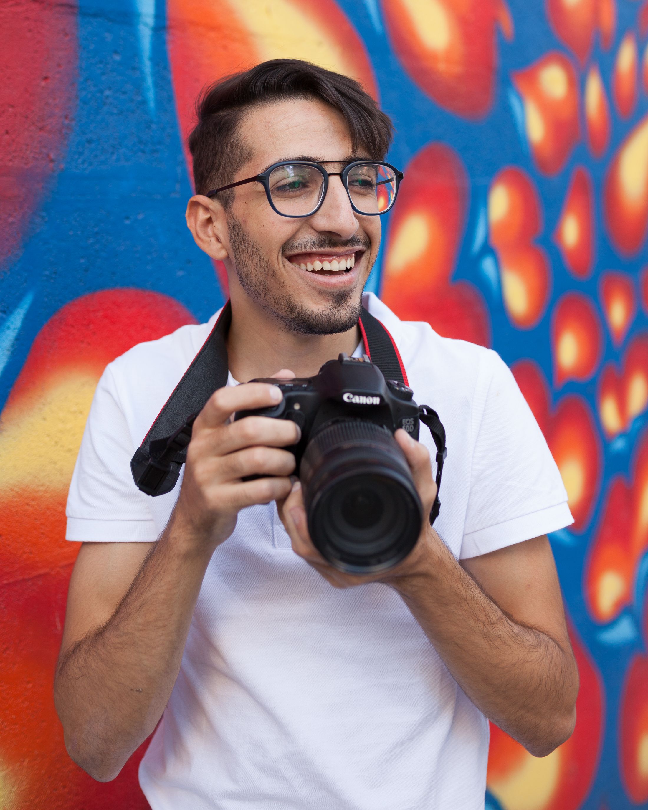 Young male photographer with a camera smiles against a colorful mural background.