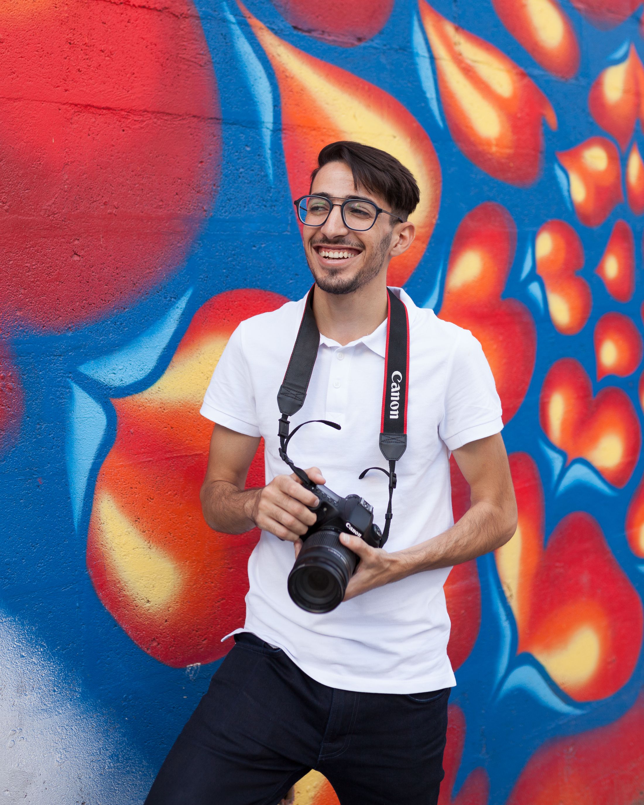 Urban portrait of a smiling man with a Canon camera, standing by a vibrant mural.