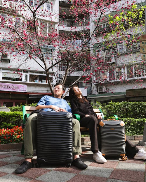 A Man and A Woman Resting On the Bench with Their Travel Cases Under an Apartment Building