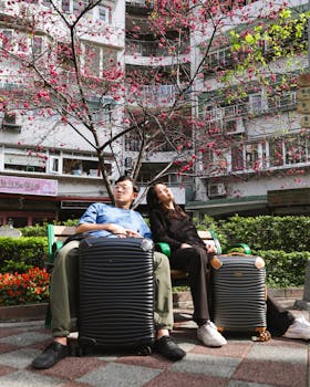 Couple resting with luggage under cherry blossoms in urban park, vibrant spring scene.