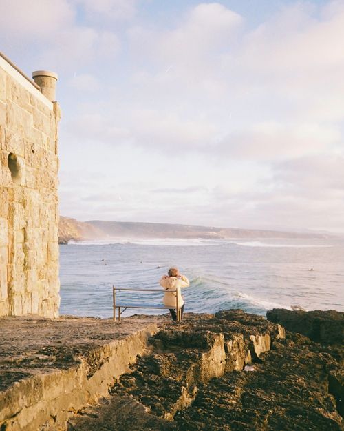 Free A woman sitting on a bench by the ocean in Ericeira, Lisbon, enjoying a serene coastal view. Stock Photo