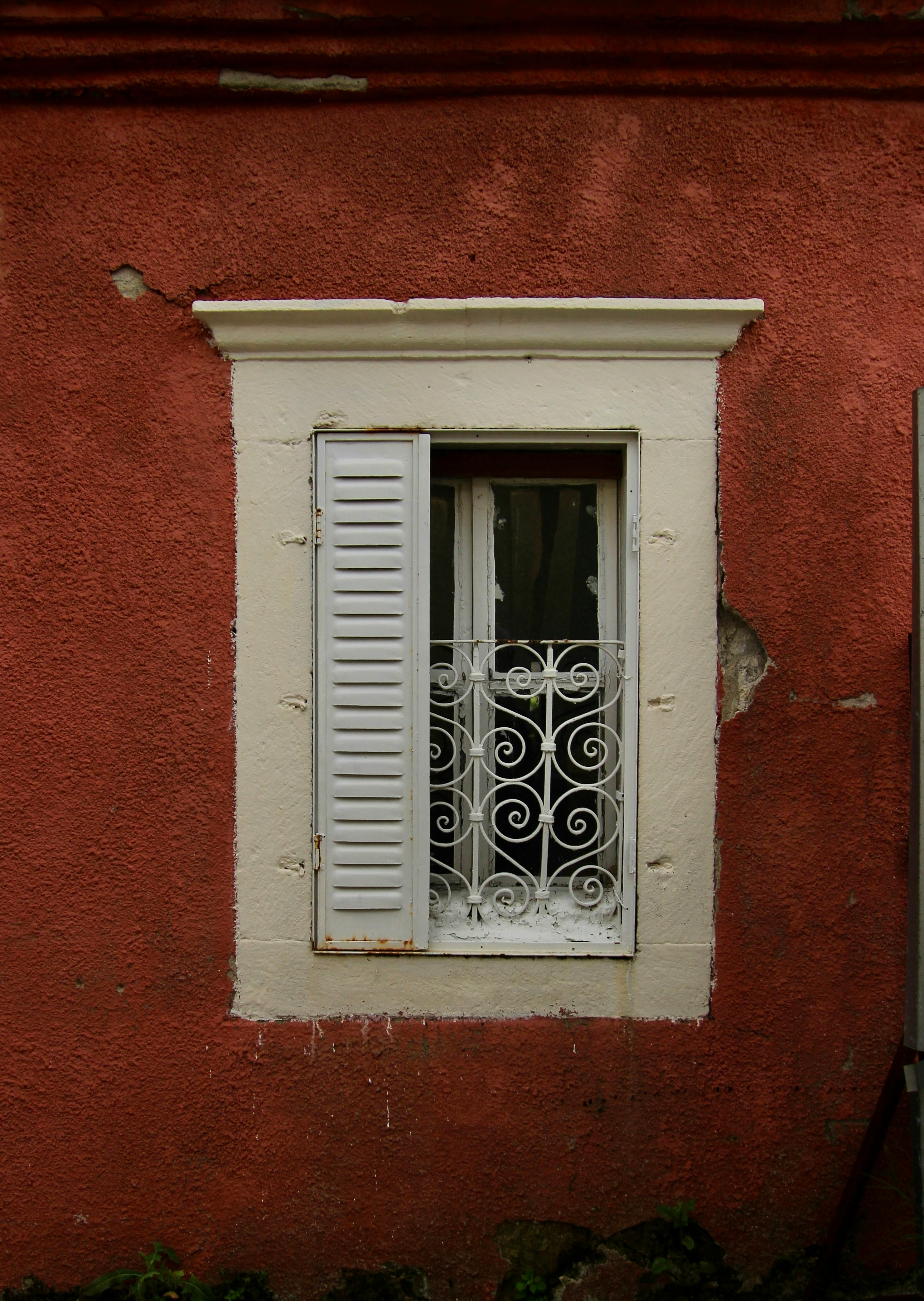 Weathered red textured wall featuring a classic white window with partially open shutters and ornate metal grilles.