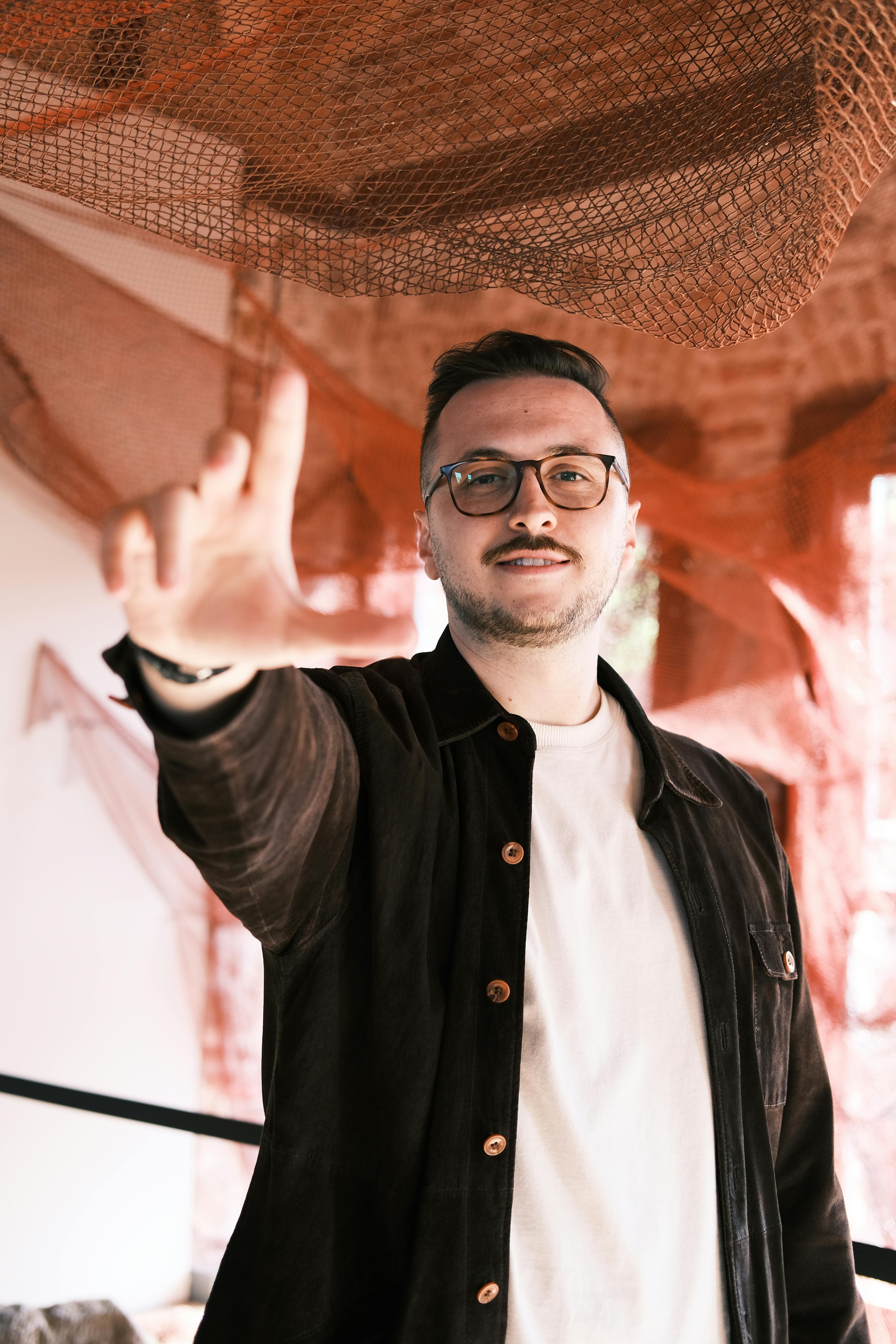 A man posing indoors with red artistic decor, showcasing a casual style.