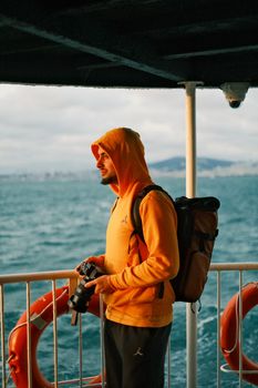 Man in orange hoodie holding camera, standing on ferry with ocean background.