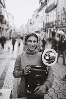 Smiling woman holding a vintage camera on a busy city street, capturing moments in black and white.