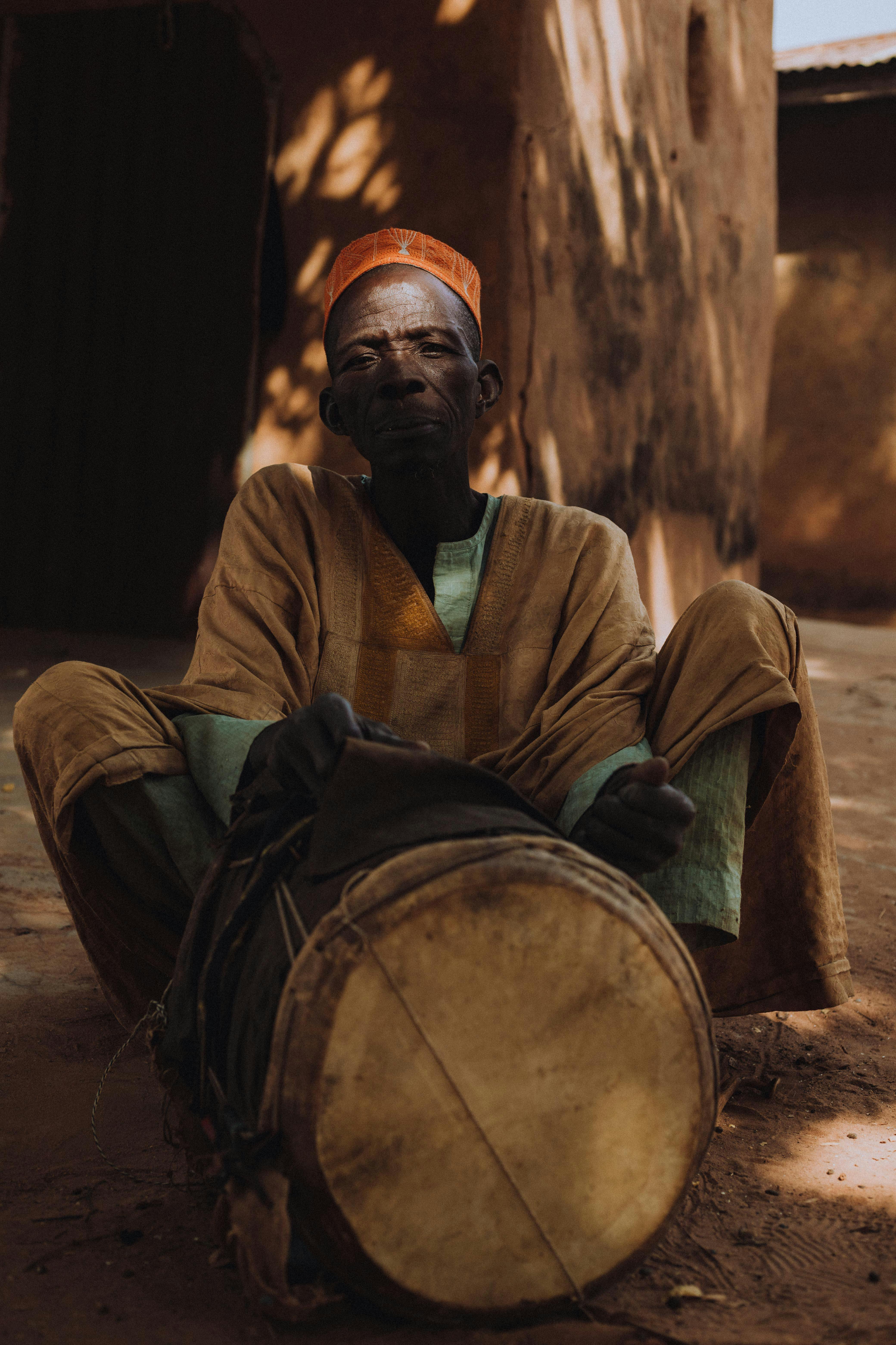 An elderly man in traditional clothing sitting on the ground playing a drum outdoors.