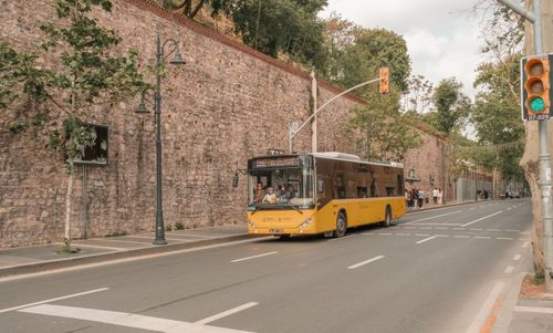 Free A yellow city bus travels along an urban street near a historic stone wall and trees. Stock Photo
