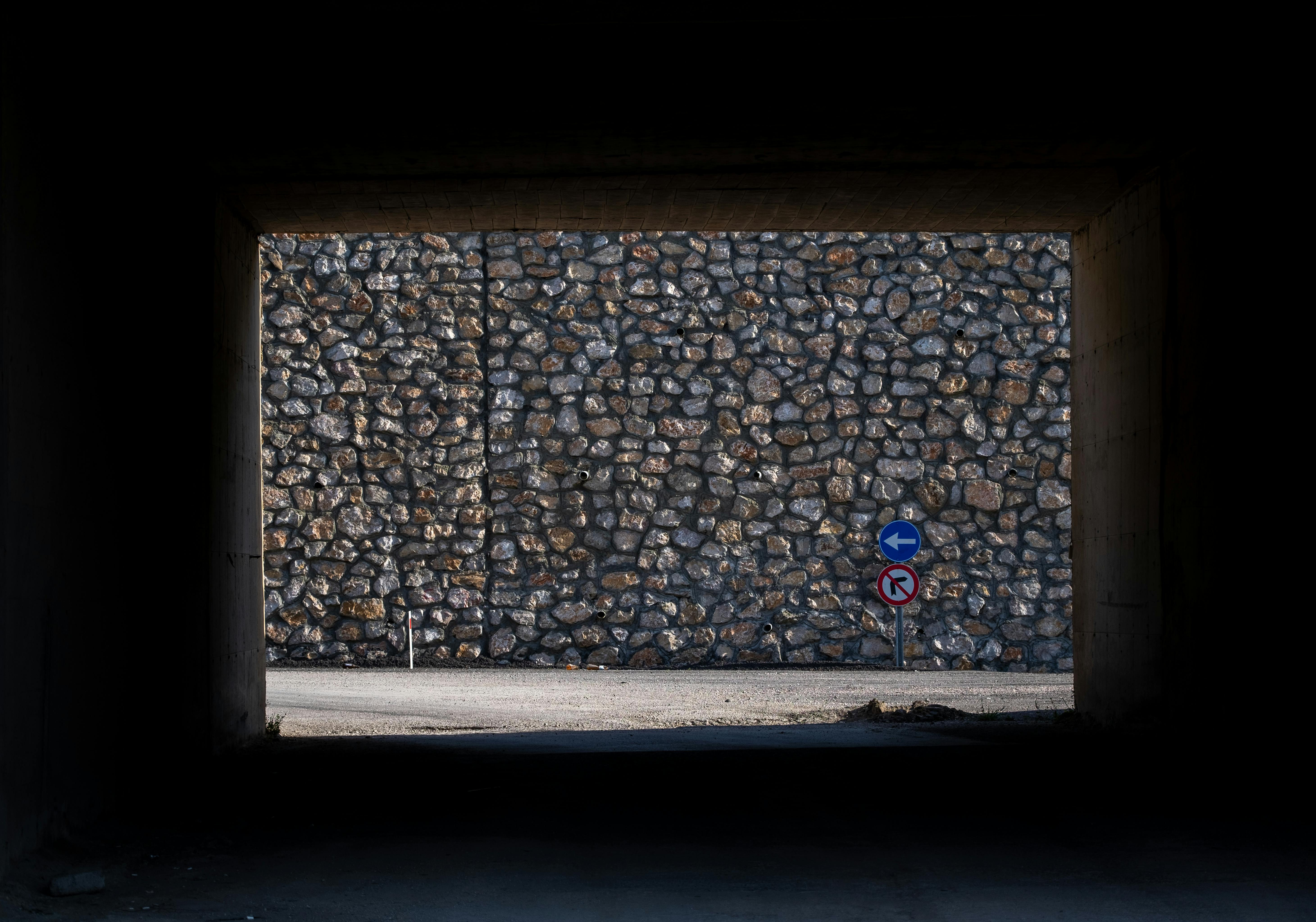 A shadowy tunnel frames a stone wall with road signs, creating an urban scene.