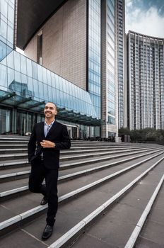Smiling businessman in a black suit walks confidently by a modern office building in urban setting.