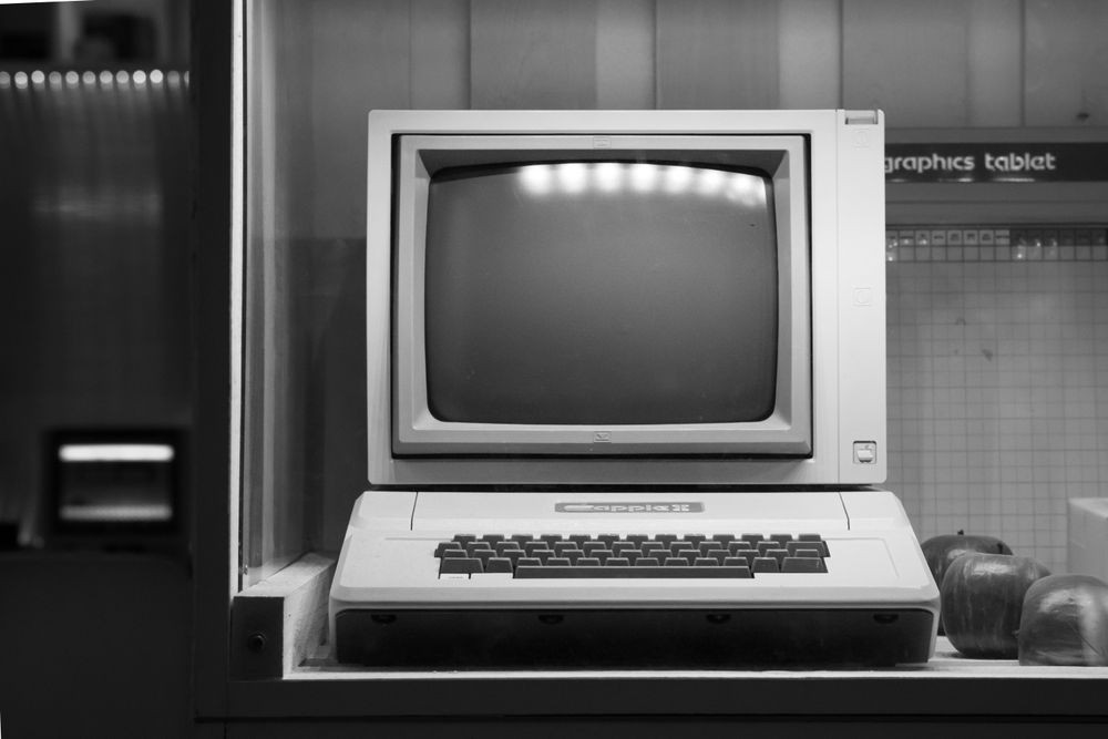 A black and white photograph of an early desktop computer terminal, boxy monitor and keyboard, standing alone against a bare institutional wall