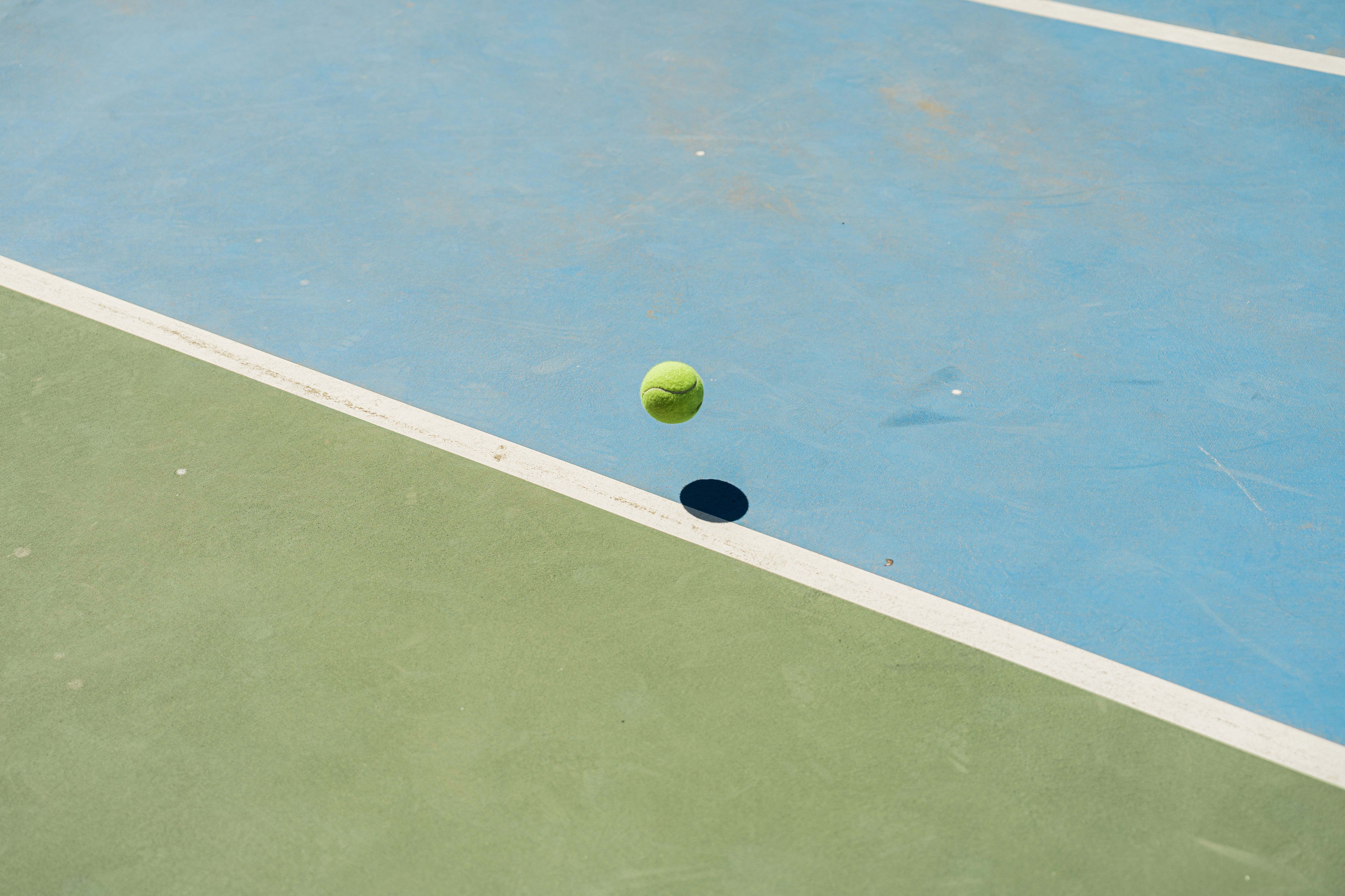 A tennis ball bounces midair creating a shadow on a blue and green tennis court.