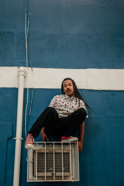 Man with dreadlocks posing on outdoor air conditioner in urban setting.