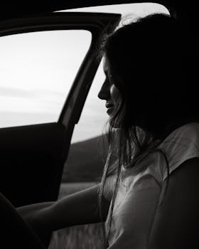 Black and white image of a woman sitting in a car doorway, reflecting twilight mood.