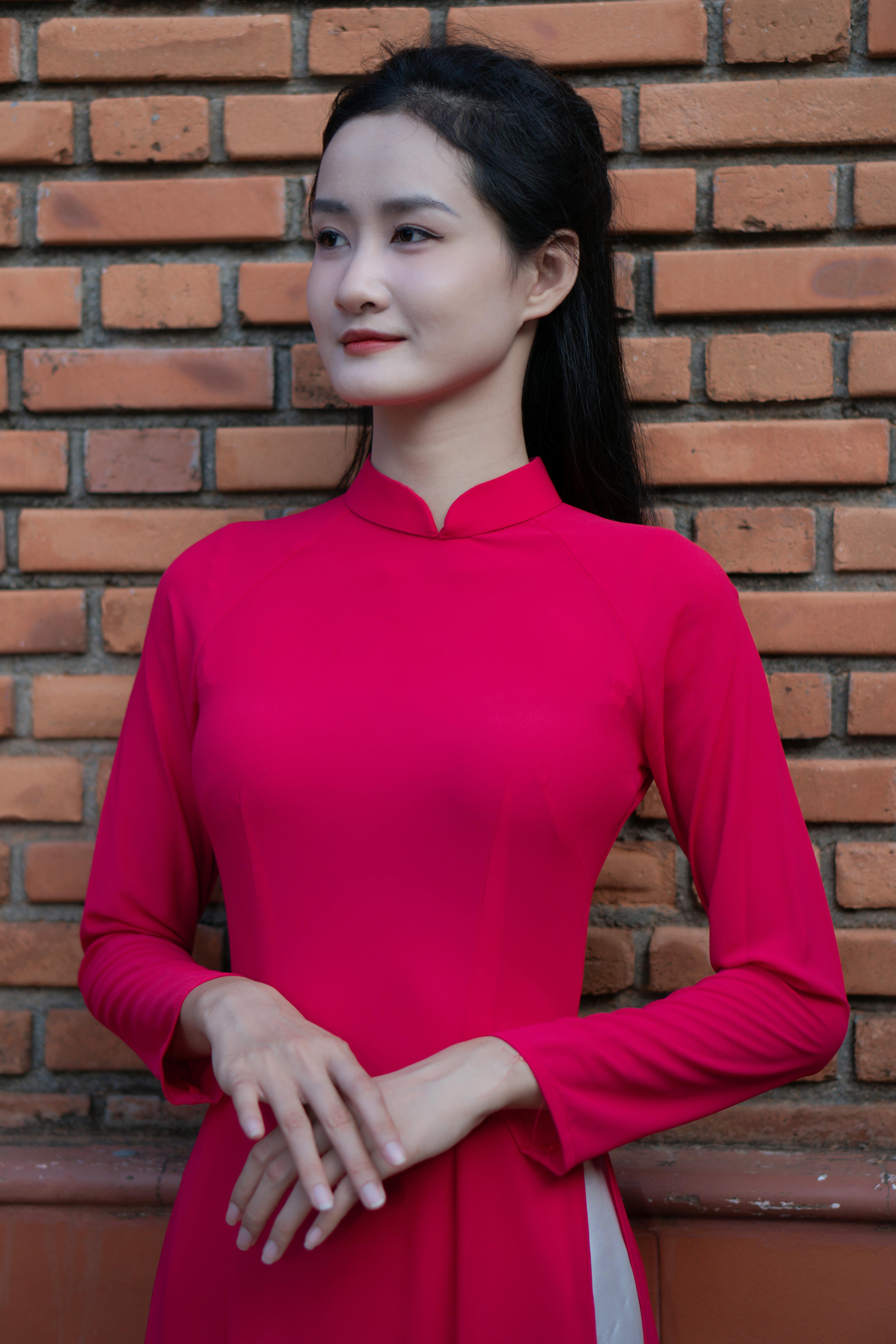 Portrait of an Asian woman in a red ao dai standing by a brick wall.