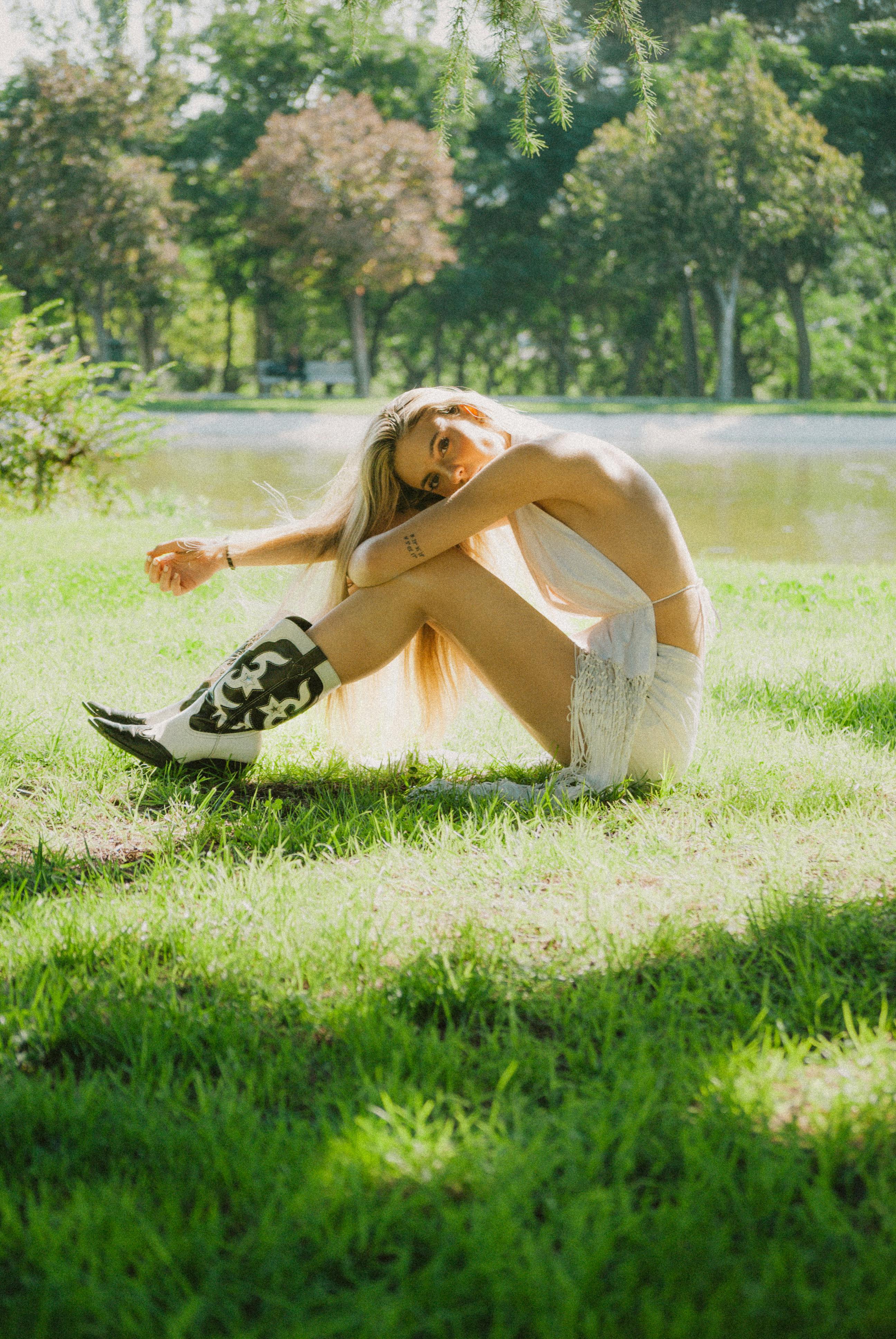 A woman in a white dress and cowboy boots sits relaxed on green grass in a sunny park.