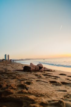 Woman in bikini relaxing on a sandy beach during sunset with city skyline in the distance.