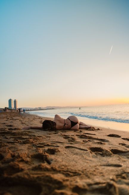 Woman in bikini relaxing on a sandy beach during sunset with city skyline in the distance.